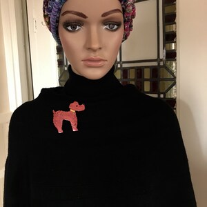 May include: A mannequin wearing a black turtleneck sweater and a colorful knit hat. A red poodle brooch with a yellow collar is pinned on the sweater. The hat features a mix of colors including pink, blue, and green.