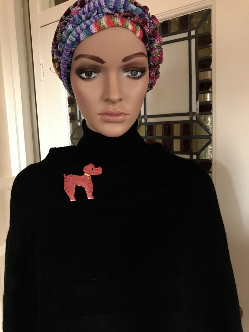 May include: A mannequin wearing a black turtleneck sweater and a colorful knit hat. A red poodle brooch with a yellow collar is pinned on the sweater. The hat features a mix of colors including pink, blue, and green.