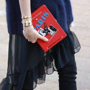 May include: A rectangular red book-shaped clutch bag featuring the word PARIS in blue letters, a black Eiffel Tower graphic, and white text. The accessory is held alongside two gold-toned metal cuff bracelets with engraved details.