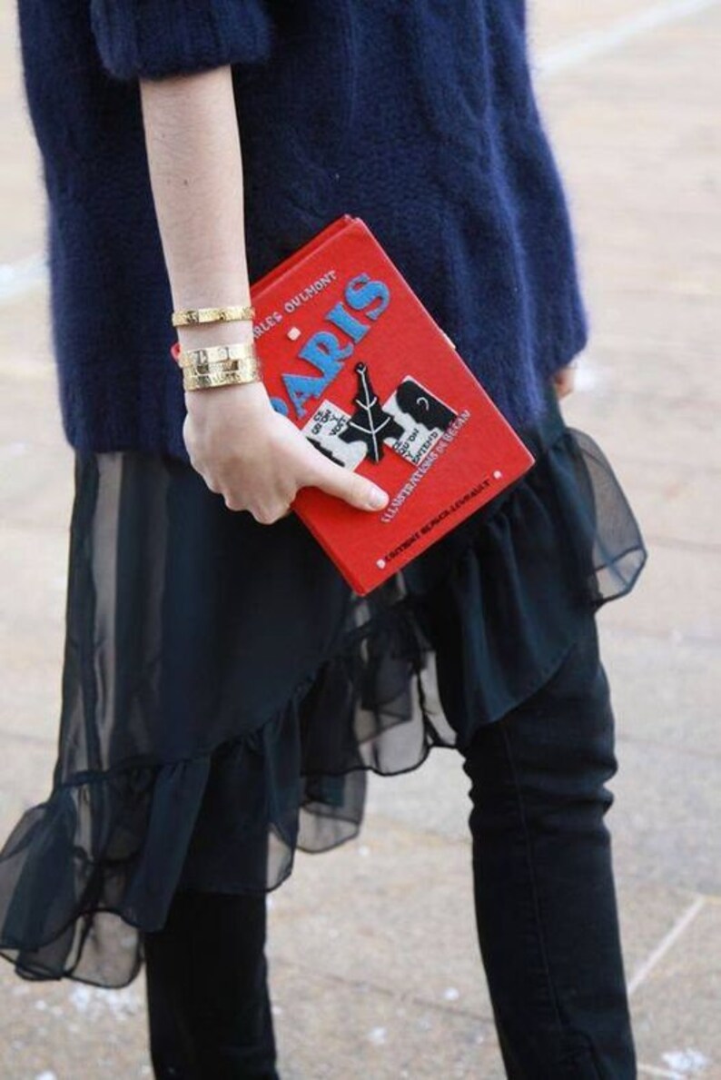 May include: A rectangular red book-shaped clutch bag featuring the word PARIS in blue letters, a black Eiffel Tower graphic, and white text. The accessory is held alongside two gold-toned metal cuff bracelets with engraved details.