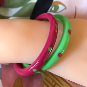 May include: Two bangles, one magenta and one green, are stacked on a wrist. The magenta bangle has green polka dots, and the green bangle has red heart-shaped accents. The bangles are made of a glossy material.