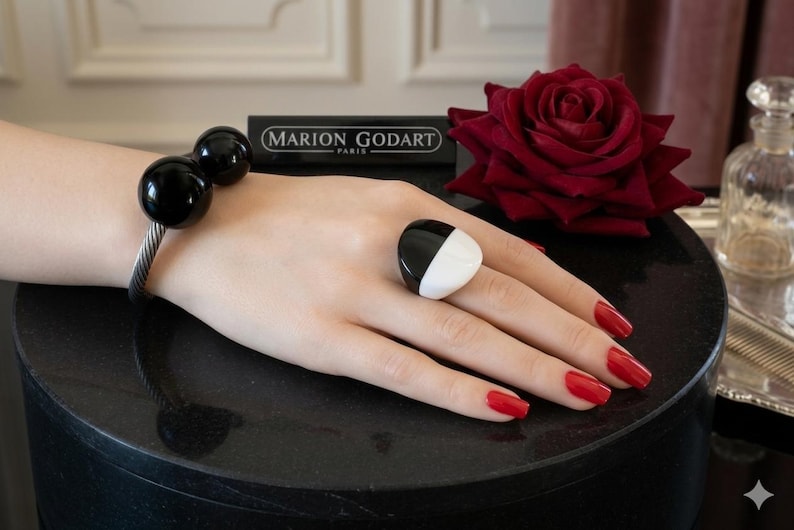 May include: A black and white ring and a bracelet with two black spheres are displayed. The ring has a geometric design. The bracelet is made of a silver-colored metal. The nails are painted red. The brand name "MARION GODART" is visible.