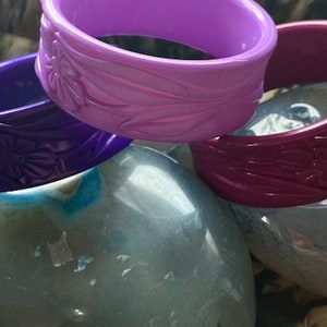 May include: Three plastic bangles with floral designs. The bangles are purple, pink, and burgundy.