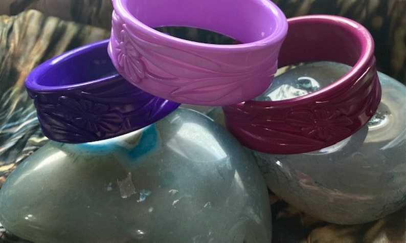 May include: Three plastic bangles with floral designs. The bangles are purple, pink, and burgundy.