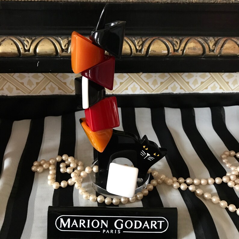 May include: A black cat-shaped ring holder with a stack of colorful rings in orange, red, black, and white. The holder has a white square base and the brand name "MARION GODART PARIS" is visible.