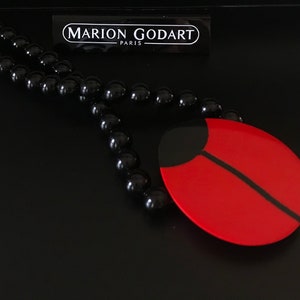 May include: A black beaded necklace with a red and black ladybug pendant. The pendant is made of resin and has a glossy finish. The necklace is from the brand "MARION GODART - PARIS".
