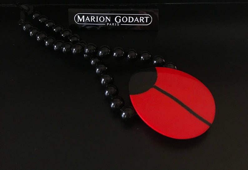 May include: A black beaded necklace with a red and black ladybug pendant. The pendant is made of resin and has a glossy finish. The necklace is from the brand "MARION GODART - PARIS".