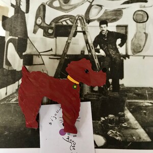 May include: A wooden dog-shaped brooch in shades of red with a yellow collar and green accent. The brooch is set against a black and white photograph of an artist's studio. The text "Pavone Paris" is visible.