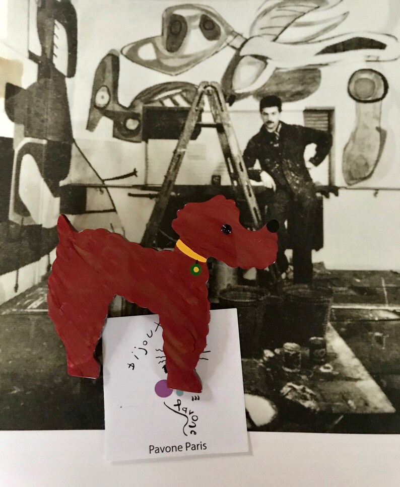 May include: A wooden dog-shaped brooch in shades of red with a yellow collar and green accent. The brooch is set against a black and white photograph of an artist's studio. The text "Pavone Paris" is visible.