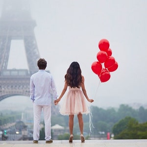 May include: A couple holding hands in front of the Eiffel Tower in Paris, France. The woman is holding a bunch of red balloons.