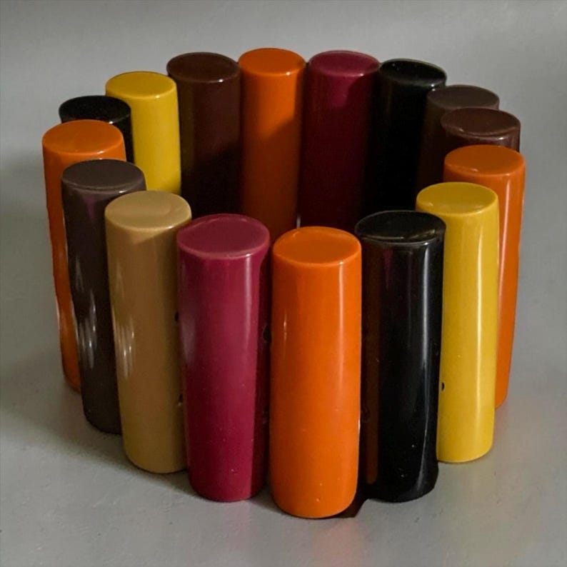 May include: A circular bracelet composed of multiple vertical, cylindrical segments in a variety of colours including orange, yellow, brown, black, and deep magenta. The segments are arranged side-by-side to form a continuous, flexible band.