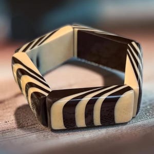 May include: A rigid bracelet composed of six rectangular segments connected to form a hexagonal shape. Each segment features a high-contrast pattern of alternating smooth black and cream-colored curved stripes that wrap around the surface.