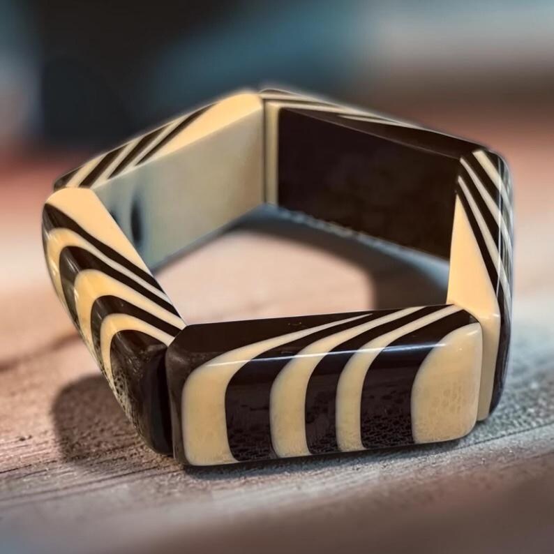 May include: A rigid bracelet composed of six rectangular segments connected to form a hexagonal shape. Each segment features a high-contrast pattern of alternating smooth black and cream-colored curved stripes that wrap around the surface.