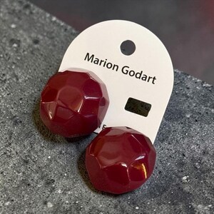May include: A pair of dark red, faceted, round stud earrings attached to a white display card featuring the text Marion Godart. The earrings have a glossy, multi-faceted surface that reflects light, creating a geometric appearance on each piece.