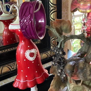 May include: A purple plastic bangle with a floral design, displayed on a white mannequin wearing a red polka dot dress with a white heart on the front. The mannequin is on a white stand with a red bow.