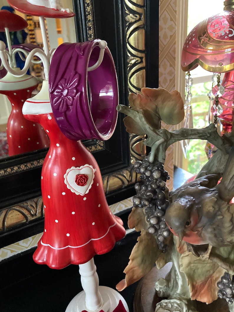 May include: A purple plastic bangle with a floral design, displayed on a white mannequin wearing a red polka dot dress with a white heart on the front. The mannequin is on a white stand with a red bow.