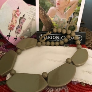 May include: A taupe statement necklace featuring large, geometric shapes and smaller round beads. The necklace is displayed on a white surface, with a black and white card that reads "MARION GODART PARIS".