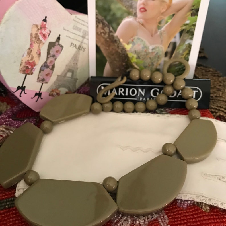 May include: A taupe statement necklace featuring large, geometric shapes and smaller round beads. The necklace is displayed on a white surface, with a black and white card that reads "MARION GODART PARIS".