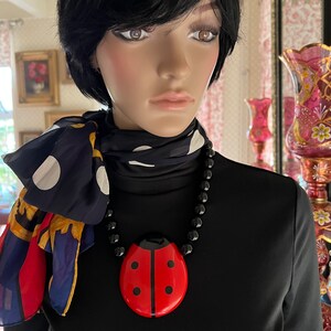 May include: A black necklace with a large red ladybug pendant. The necklace is worn by a mannequin with black hair and a black and white polka dot scarf.