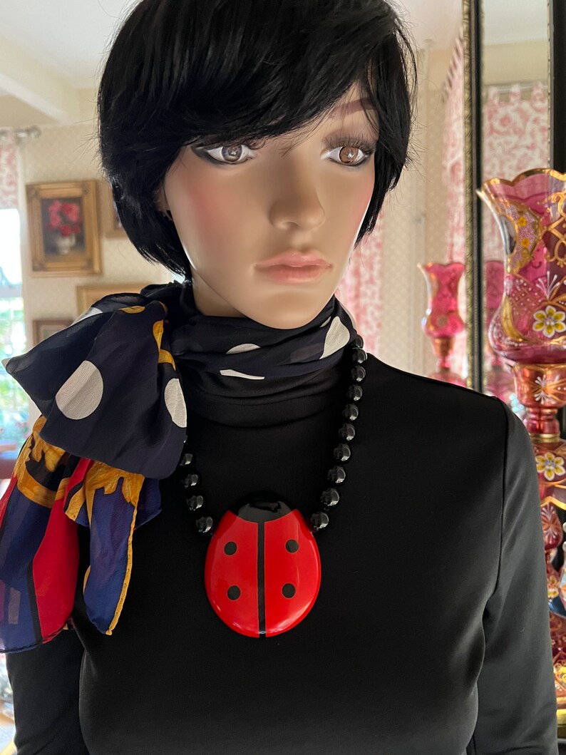 May include: A black necklace with a large red ladybug pendant. The necklace is worn by a mannequin with black hair and a black and white polka dot scarf.