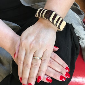 May include: A chunky, rectangular-link bracelet featuring alternating glossy dark brown and cream-colored segments. The bracelet is worn on the wrist, paired with a thin, gold-toned band ring featuring a row of small, clear, round-cut stones.