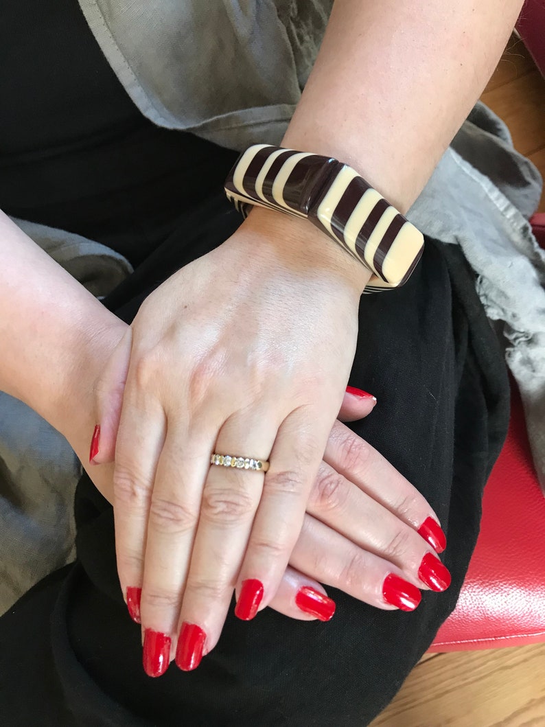 May include: A chunky, rectangular-link bracelet featuring alternating glossy dark brown and cream-colored segments. The bracelet is worn on the wrist, paired with a thin, gold-toned band ring featuring a row of small, clear, round-cut stones.