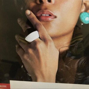 May include: A white and black ring is worn on a hand. The ring has a curved, rectangular shape. A turquoise earring is visible. The image is a close-up of a person's face and jewelry.