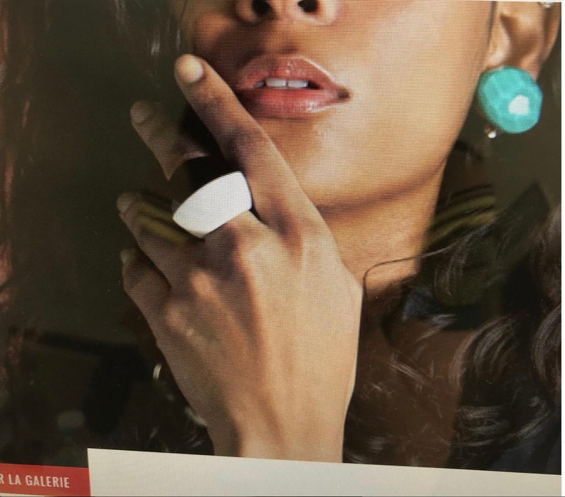 May include: A white and black ring is worn on a hand. The ring has a curved, rectangular shape. A turquoise earring is visible. The image is a close-up of a person's face and jewelry.