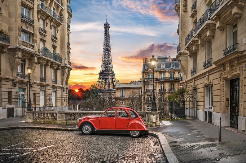 May include: A bright red vintage Citro&euml;n 2CV car parked on a cobblestone street in Paris. The Eiffel Tower is visible in the background under a sunset sky, framed by traditional stone apartment buildings with balconies and street lamps.