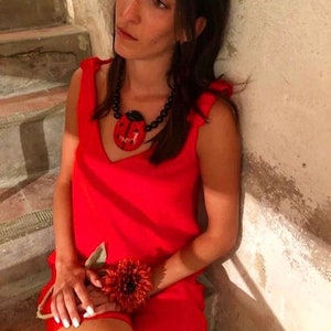 May include: A woman wearing a red sleeveless dress with a black and red ladybug pendant necklace. She is sitting on a set of stairs and holding a red flower.