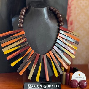 May include: A Marion Godart necklace featuring a strand of dark brown beads and a series of radiating, rectangular resin bars in shades of orange, yellow, burgundy, and brown. A matching bracelet and two round stud earrings are displayed nearby.