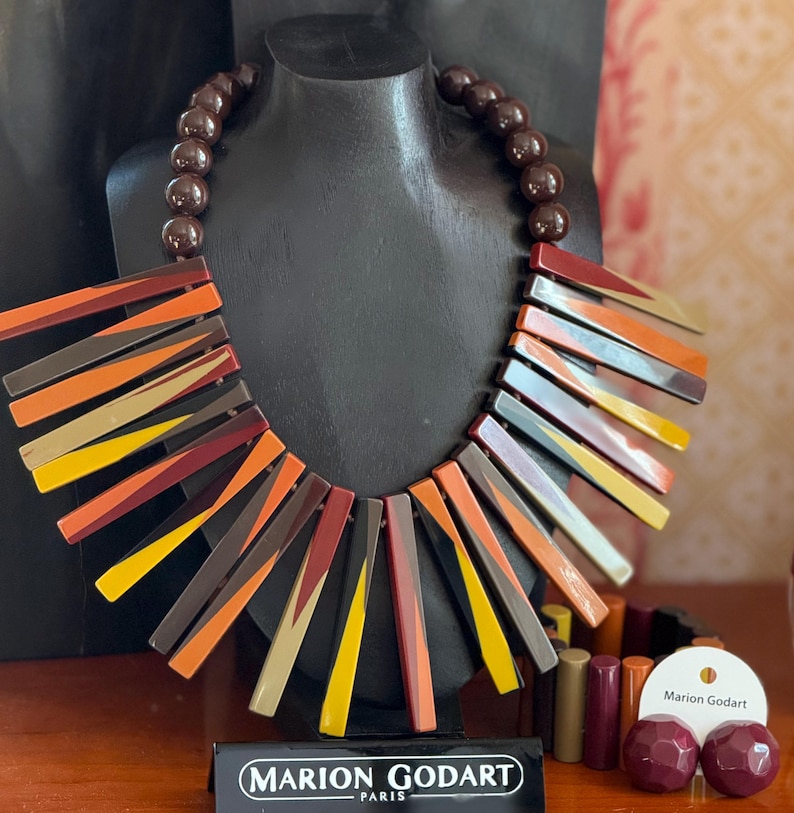 May include: A Marion Godart necklace featuring a strand of dark brown beads and a series of radiating, rectangular resin bars in shades of orange, yellow, burgundy, and brown. A matching bracelet and two round stud earrings are displayed nearby.
