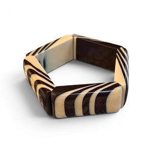 May include: A hexagonal bangle bracelet featuring alternating segments of solid dark brown and cream-colored sections with a swirling, striped pattern. The bracelet is constructed from rigid, smooth-finished pieces joined together to form a geometric shape.