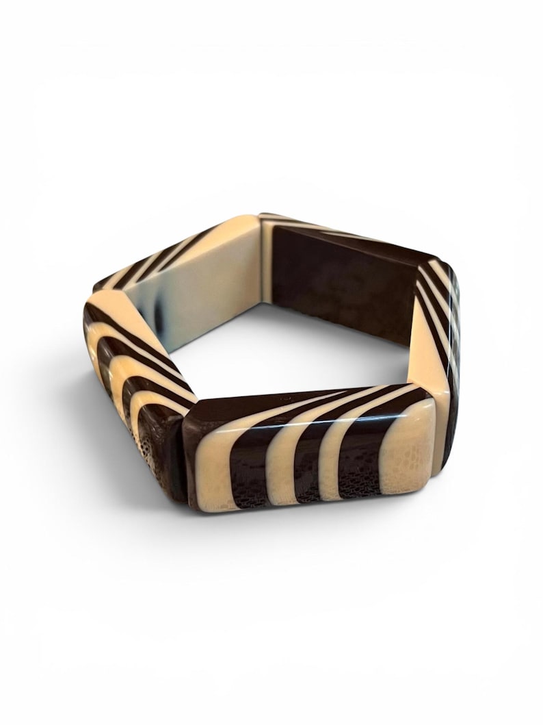 May include: A hexagonal bangle bracelet featuring alternating segments of solid dark brown and cream-colored sections with a swirling, striped pattern. The bracelet is constructed from rigid, smooth-finished pieces joined together to form a geometric shape.