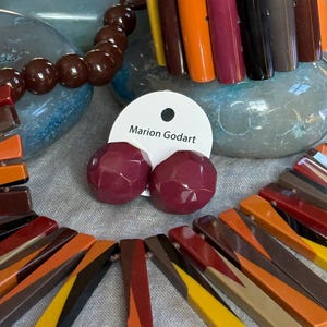 May include: A set of jewellery featuring a necklace and bracelet made of multi-coloured rectangular resin bars in shades of yellow, orange, brown, and burgundy, alongside a pair of round, faceted burgundy stud earrings on a Marion Godart card.