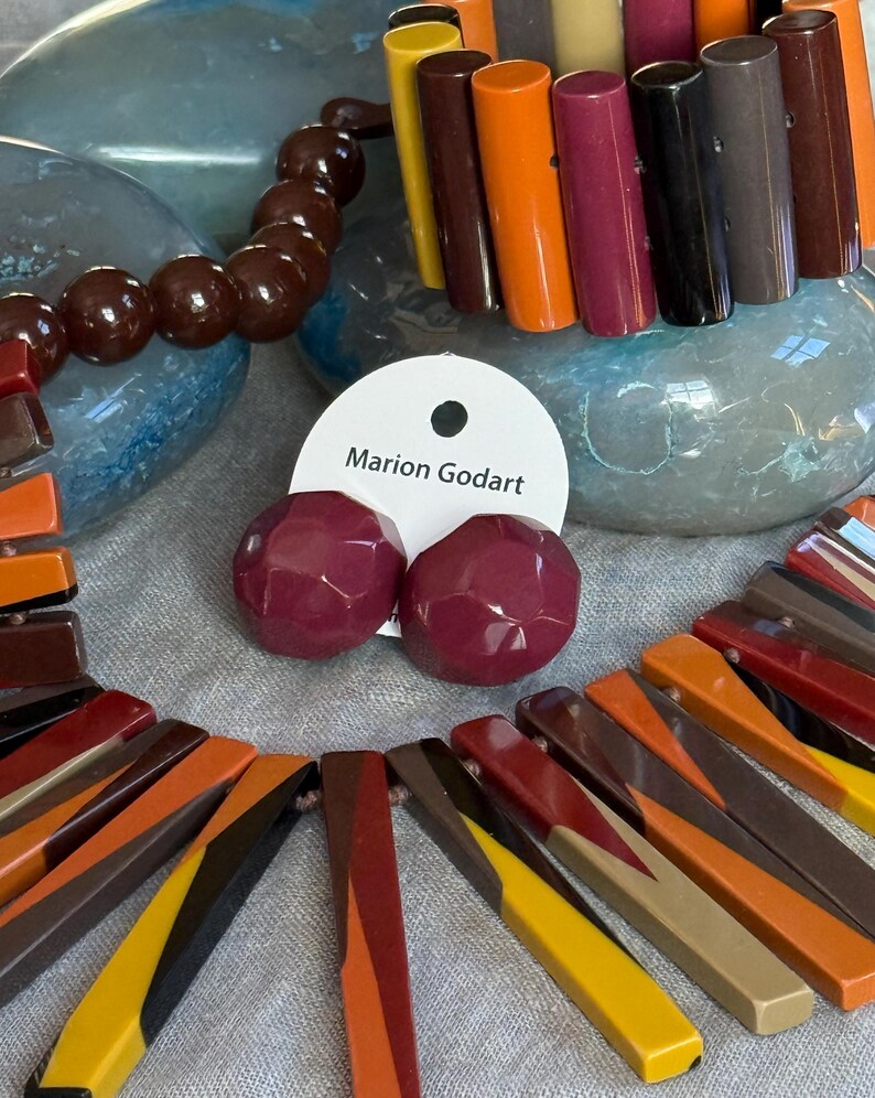 May include: A set of jewellery featuring a necklace and bracelet made of multi-coloured rectangular resin bars in shades of yellow, orange, brown, and burgundy, alongside a pair of round, faceted burgundy stud earrings on a Marion Godart card.