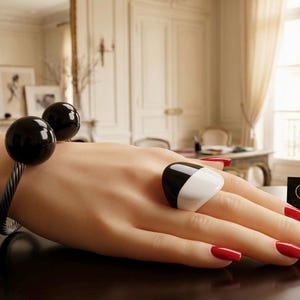 May include: A black and white ring and a bracelet with two black spheres. The hand has red nail polish. A sign that says "MARION GODART PARIS" is in the background. The jewelry is on a dark surface.