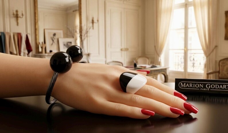 May include: A black and white ring and a bracelet with two black spheres. The hand has red nail polish. A sign that says "MARION GODART PARIS" is in the background. The jewelry is on a dark surface.