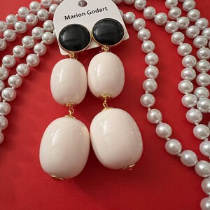 Puede incluir: Un par de pendientes de declaración blancos y negros con un acento dorado. Los pendientes están hechos de dos cuentas ovaladas blancas grandes y una cuenta redonda negra más pequeña. Los pendientes se muestran en una tarjeta blanca con el texto "Marion Godart" y están rodeados por un collar de perlas.