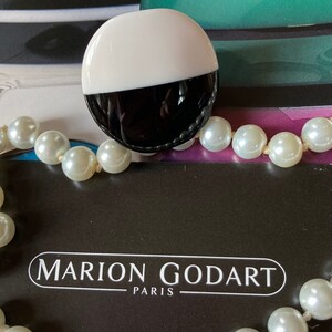 May include: A two-tone, black and white, round brooch with a pearl necklace. The brooch is half white and half black. The necklace has large, round, white pearls. The text "MARION GODART PARIS" is visible.