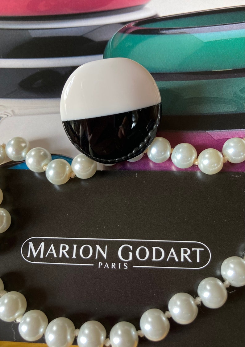 May include: A two-tone, black and white, round brooch with a pearl necklace. The brooch is half white and half black. The necklace has large, round, white pearls. The text "MARION GODART PARIS" is visible.