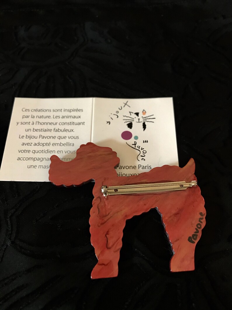 May include: A red and brown poodle-shaped brooch with a silver pin. The brooch has a marbled effect and is set against a dark background. A small card with French text is visible.
