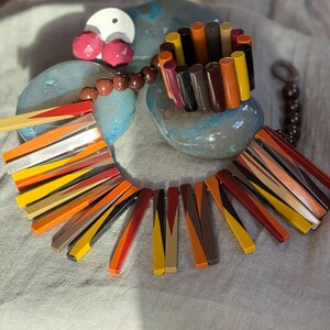 May include: A statement necklace and matching bracelet set featuring cylindrical beads in a palette of red, orange, yellow, brown, and black. The necklace includes a strand of brown spherical beads and a series of long, flat, multi-coloured rectangular segments arranged in a fan-like pattern.