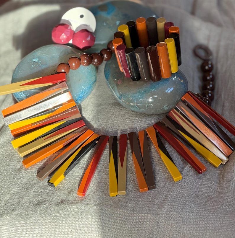 May include: A statement necklace and matching bracelet set featuring cylindrical beads in a palette of red, orange, yellow, brown, and black. The necklace includes a strand of brown spherical beads and a series of long, flat, multi-coloured rectangular segments arranged in a fan-like pattern.