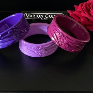 May include: Three purple plastic bangles with floral designs. The bangles are arranged in a gradient from dark purple to light purple. The text "MARION GOD PARIS" is visible in the background.