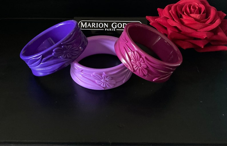 May include: Three purple plastic bangles with floral designs. The bangles are arranged in a gradient from dark purple to light purple. The text "MARION GOD PARIS" is visible in the background.