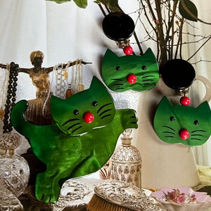 May include: Three green cat-shaped earrings with black and red details. The earrings are made of a shiny, green material and have a black button on the top. The earrings are hanging from a branch with a white background.