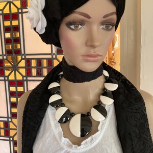 May include: A mannequin wearing a black and white statement necklace with large, circular beads. The necklace is paired with a black turban adorned with a white flower and a black scarf. The mannequin is wearing a white top.