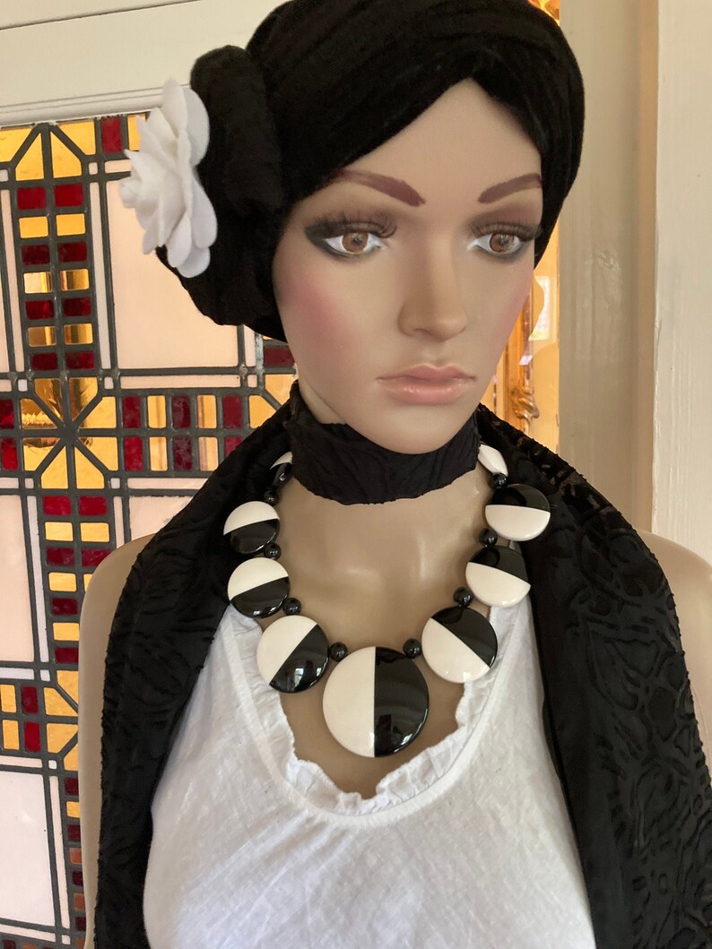 May include: A mannequin wearing a black and white statement necklace with large, circular beads. The necklace is paired with a black turban adorned with a white flower and a black scarf. The mannequin is wearing a white top.