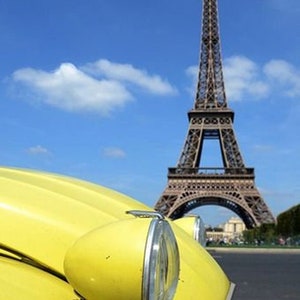 May include: A yellow vintage car with a large headlight is in the foreground, with the Eiffel Tower in the background under a blue sky with clouds. The car's bonnet curves towards the camera.